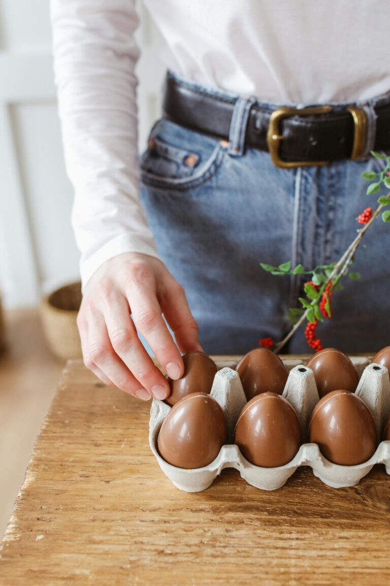 A person arranging chocolate eggs in a carton on a wooden table, indoors.
